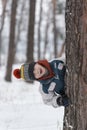 Cheerful boy peeks out from behind tree. Child in knitted hat walks through winter snow-kept park Royalty Free Stock Photo