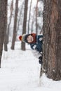 Cheerful boy peeks out from behind tree. Child in knitted hat walks through winter snow-kept park. Vertical frame Royalty Free Stock Photo