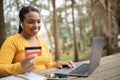 Cheerful brazilian lady holding credit card, typing on laptop, enjoying online sales, sitting in city park, free space Royalty Free Stock Photo