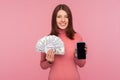 Cheerful brunette woman in pink sweater holding dollars banknotes and cell phone with mock up blank display, online payments, Royalty Free Stock Photo