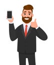Cheerful businessman holding/showing brand new smartphone/mobile/cell phone in hand and gesturing thumbs up sign. Good, deal.