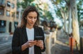 Cheerful businesswoman in jacket with cell phone in park, portrait elegant woman with smartphone and wireless earphones on street