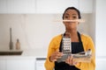 Cheerful busy funny millennial african american woman chef in apron carries kitchenware and spoon under her nose