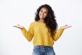 Cheerful carefree alluring african-american woman with curly dark hairstyle in yellow sweater, spread hands sideways
