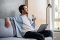 Cheerful caucasian man wearing casual clothes sitting on the sofa and using laptop at home. Inspired male entrepreneur