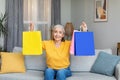 Cheerful caucasian mature gray-haired woman shows colored packages with purchases, sits on sofa Royalty Free Stock Photo