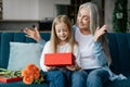 Cheerful caucasian small girl opens gift box with retired granny with bouquet of flowers and enjoys surprise