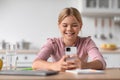 Cheerful caucasian teen girl student chatting at phone, sits at table in kitchen interior