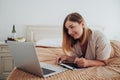 Cheerful Caucasian Woman Laying on the Bed in Hotel Room and Working on a Digital Tablet and Laptop, Designer Freelancer