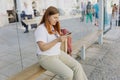 A cheerful caucasian woman is sitting on a wooden bench inside of an urban bus stop and using paper map. Urban travel Royalty Free Stock Photo