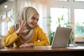 Cheerful Asian-Muslim woman is greeting her colleague via video call or online meeting on her laptop Royalty Free Stock Photo