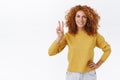 Cheerful, confident redhead curly woman in yellow sweater showing peace, goodwill sign and smiling determined, hold arm