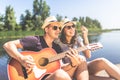 Cheerful couple with acoustic guitar sitting on pier and singing. Lake and trees in the background