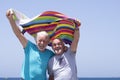 Cheerful couple of senior bearded brothers laughing at the beach holding a colorful towel in the wind - concept of freedom and Royalty Free Stock Photo