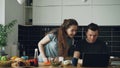 Cheerful couple at table woman dancing and cooking while gives hasband to try red pepper, man sitting working on laptop