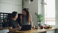 Cheerful couple at table woman dancing and cooking while gives hasband to try red pepper, man sitting working on laptop