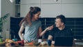 Cheerful couple at table woman cooking while gives hasband to try red pepper, man sitting working on laptop