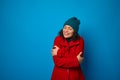 Cheerful curly-haired brunette pretty woman dressed in bright red coat and warm woolen green hat hugging warming herself poses