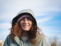 Cheerful Curly-Haired Woman in Hood and Glasses Against Blue Sky Royalty Free Stock Photo