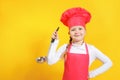 Cheerful cute baby chef holds soup ladle in hands. Little girl in uniform on a yellow background