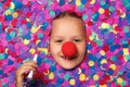 Cheerful cute face of a little girl with a red nose in colorful confetti. The child holds a whistle in his hand Royalty Free Stock Photo