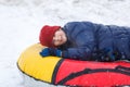 Cheerful cute young boy in orange hat red scarf and blue jacket holds tube on snow, has fun, smiles. Teenager on sledding Royalty Free Stock Photo