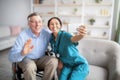 Cheerful disabled senior man in wheelchair and his nurse taking selfie indoors, selective focus Royalty Free Stock Photo