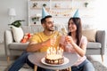 Cheerful diverse couple in festive hats toasting with champagne glasses, having birthday cake with candles at home Royalty Free Stock Photo