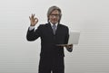 Cheerful elderly asian businessman in black suit and eyeglasses holding laptop computer and showing ok gesture sign with fingers, Royalty Free Stock Photo