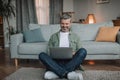 Cheerful elderly european man with beard blogger typing on pc, sits on floor in living room interior