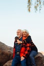 Cheerful elderly people a woman and a man are sitting on the stones and hugging on the lake, against the background of the bridge Royalty Free Stock Photo