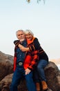 Cheerful elderly people a woman and a man are sitting on the stones and hugging on the lake, against the background of the bridge Royalty Free Stock Photo