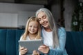 Cheerful European Little Girl And Old Woman Watch Video In Tablet, Read Book Online In Living Room Interior