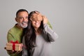 Cheerful european old husband closes eyes to his wife, gives box with gift