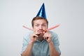 cheerful european young man having fun on party wearing blue denim shirt and holiday hat, blowing party horn