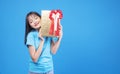 Cheerful excited asian woman holding gold gift box with red ribbon in own hand while standing over isolated blue background. Royalty Free Stock Photo