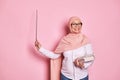 Cheerful experienced middle aged Arab Musim teacher in pink hijab, points with pointer at copy space on pink background