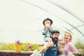 Cheerful family in greenhouse. Father in blue vest holding his son on shoulders while kid is eating apple. Bearded man