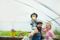 Cheerful family in greenhouse. Father in blue vest holding his son on shoulders while kid is eating apple. Bearded man