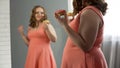 Cheerful fat girl satisfied with her appearance eating donuts in front of mirror