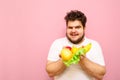 Cheerful fat man in white t-shirt stands on a pink background and holds fruits and salad leaves in his hands, looks in camera and