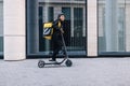 Cheerful female courier standing on electric scooter. Woman in cycling helmet wearing thermal backpack looking away