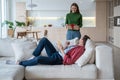 Smiling woman coming to sofa with festive giftbox, looking at resting man who doesnt expect surprise Royalty Free Stock Photo