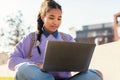 Cheerful female mixed race student with laptop computer doing homework or attending online lecture, sitting outdoors Royalty Free Stock Photo