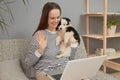 Cheerful friendly Caucasian woman wearing striped shirt sitting on sofa with her puppy dog using laptop computer having video call Royalty Free Stock Photo