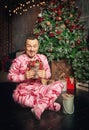 Cheerful funny man in pink sleepwear near decorated fir tree and hold Christmas present Royalty Free Stock Photo