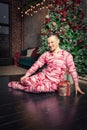 Cheerful funny man in sleepwear sitting on the floor near decorated fir tree and hold Christmas present Royalty Free Stock Photo