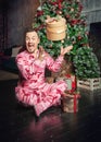 Cheerful funny man in pink sleepwear sitting near decorated fir tree and throws up Christmas present