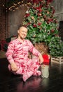 Cheerful funny man in sleepwear sitting on the floor near decorated fir tree and Christmas present Royalty Free Stock Photo