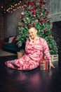 Cheerful funny man in sleepwear sitting on the floor near decorated fir tree and hold Christmas present Royalty Free Stock Photo